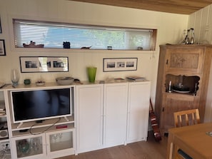 Ein kompakter Wohnbereich mit Fernseher, Stauraumschränken, Esstisch und einem Fenster mit Jalousien.