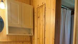 Une salle de bain aux murs lambrissés, équipée de toilettes, d’une étagère au-dessus des toilettes et d’un rideau de douche.