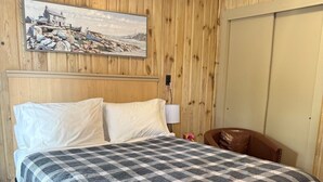 A hotel room with a wooden paneled wall, a bed with a checkered bedspread, a painting of a coastal scene, and a brown leather chair.