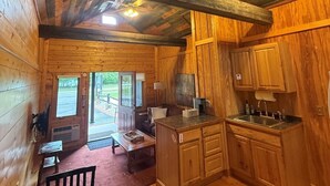 Interior de una cabaña de madera con comedor, cocina y ventana que da a una terraza exterior.