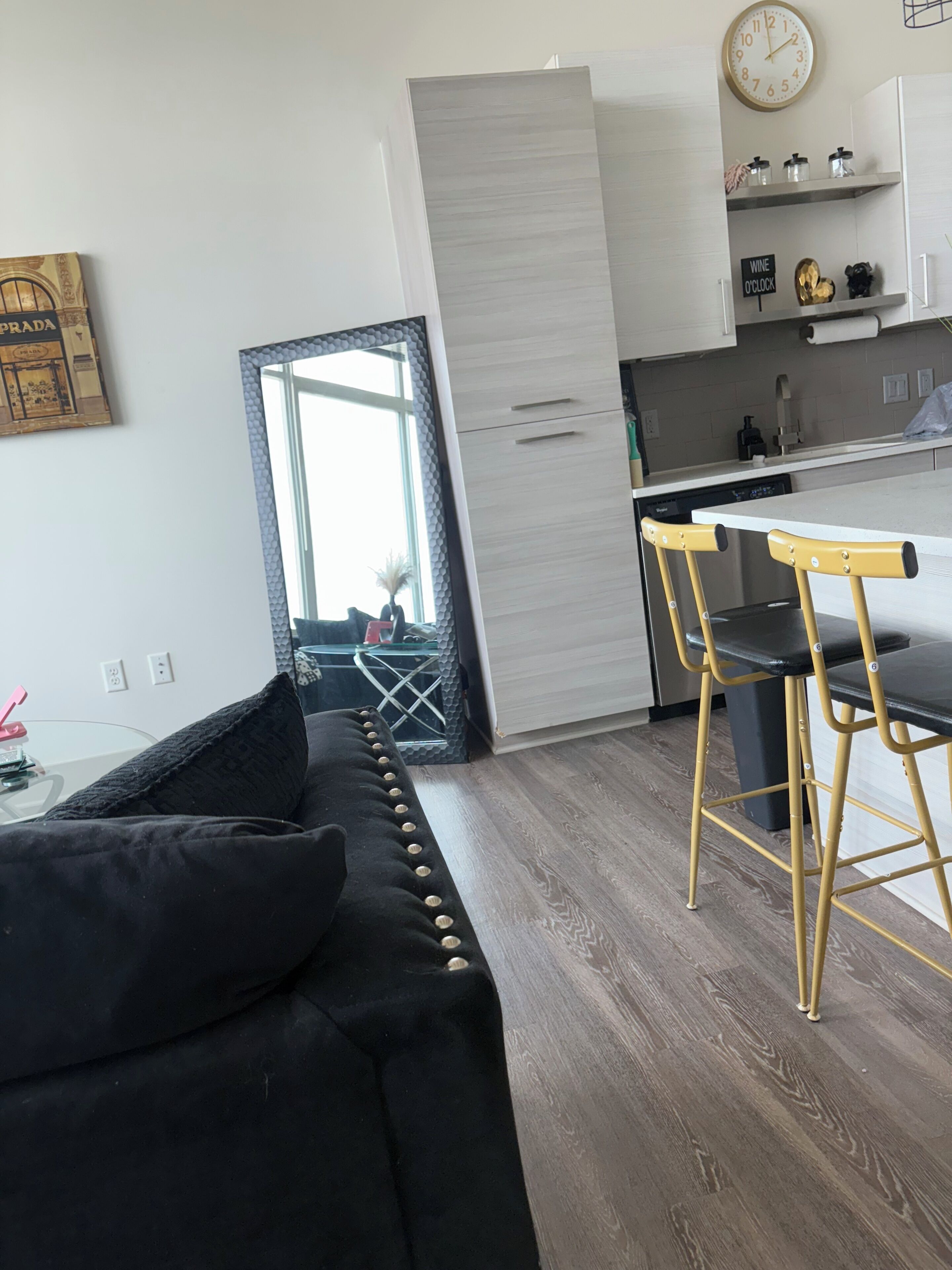 A modern kitchen with a dining area, a sofa with black cushions, and a visible clock on the wall.