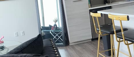 A modern kitchen with a dining area, a sofa with black cushions, and a visible clock on the wall.