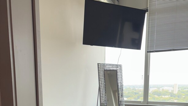 Ein Zimmer mit großem Fenster, schwarzem Teppich, Spiegel mit dekorativem Rahmen und einem an der Wand befestigten Flachbildfernseher.