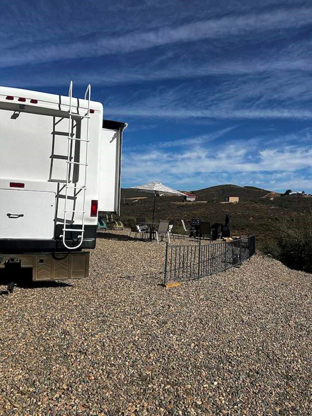A white RV parked on gravel with a ladder, a fenced area with chairs, and a mountain in the background.