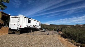 A white RV parked on a gravel driveway with a metal fence, surrounded by a rural landscape and a clear sky.