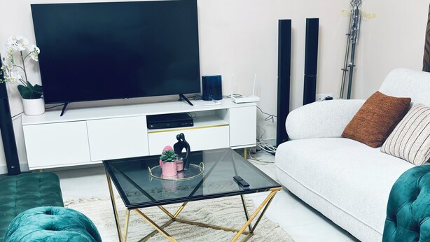 A teal upholstered sofa, a glass coffee table with a geometric gold frame, a flat-screen TV on a white cabinet, and a white sofa with a brown pillow. - Cozy apartment with a large terrace (Libreville)