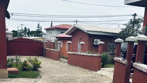 A paved courtyard with a gated entrance, surrounded by buildings with red walls and metal gates. - Cozy apartment with a large terrace (Libreville)