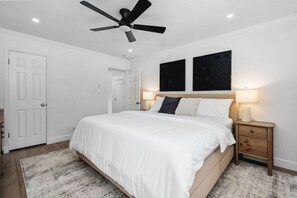 Une chambre à coucher comprenant un lit, des tables de chevet, un ventilateur de plafond et deux œuvres d’art accrochées au mur.