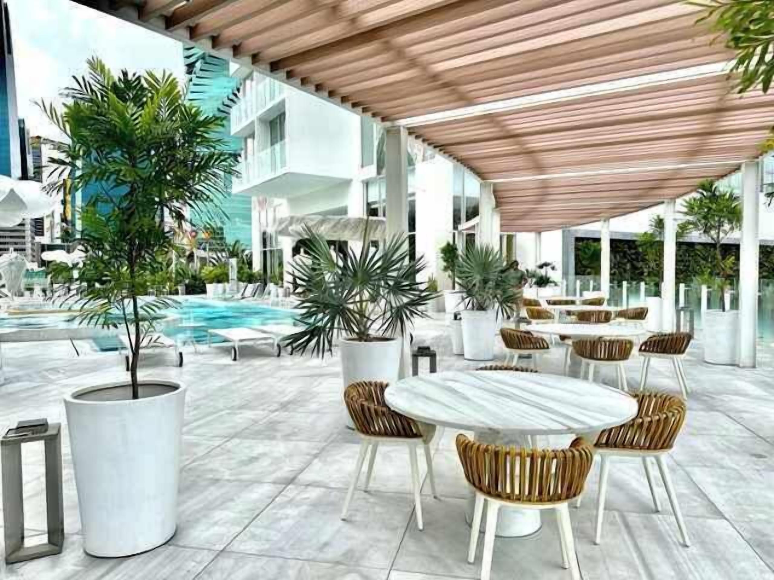 Zona de piscina al aire libre con sillones de descanso y una mesa, rodeada de plantas en macetas y arquitectura moderna.