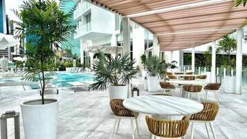 Zona de piscina al aire libre con sillones de descanso y una mesa, rodeada de plantas en macetas y arquitectura moderna.