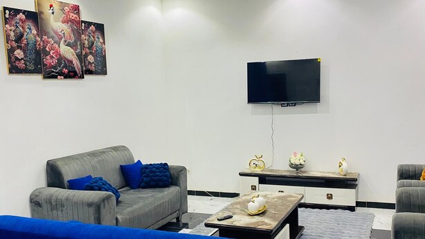 Una sala moderna con un sofá gris, un sofá azul, una mesa de centro, un televisor de pantalla plana montado en la pared, un ventilador de techo y una araña.