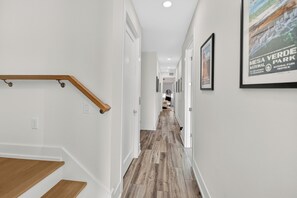 A hallway with a wooden staircase, white walls, and a wooden floor. There are framed pictures on the walls and a poster on the right side. - 52 audubon (Sarasota)