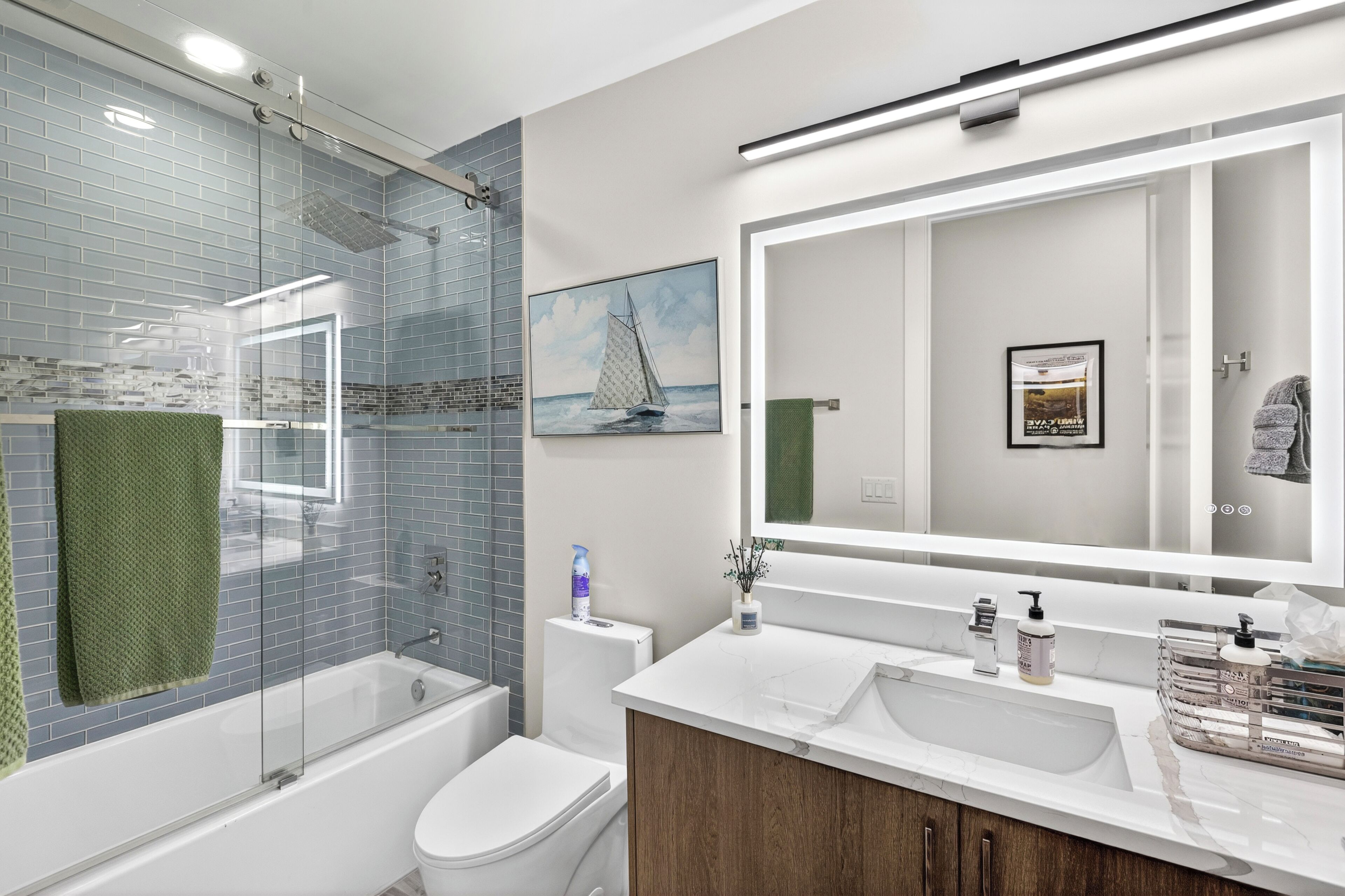 A modern bathroom with a glass shower enclosure, a toilet, a sink with a large mirror, and a framed picture on the wall.