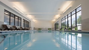 Una amplia piscina interior con agua cristalina, rodeada de sillones de descanso y grandes ventanales.