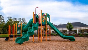 A playground with a slide and climbing structures, surrounded by trees and a building in the background. - Colonial Comfort: 3BR Lock-Off Villa with Fireplace & Full Kitchen (Williamsburg)