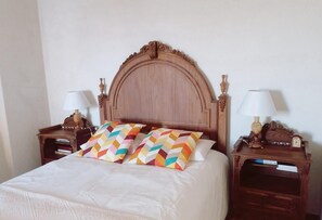 Una cama bien hecha con un cabecero de madera, dos mesitas de noche con lámparas y un cojín colorido.