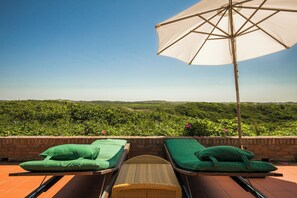 Zwei Loungestühle mit grünen Kissen, ein Korbstuhl und ein Sonnenschirm auf einer Terrasse mit Blick auf eine grüne Landschaft.