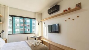 Une chambre d’hôtel comprenant un lit, une télévision, une petite table avec une chaise, une fenêtre avec des rideaux et une étagère fixée au mur, agrémentée d’objets décoratifs.