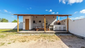 A covered outdoor seating area with a table and chairs, a wooden bench, and a small kitchen area with a stove and chairs. - House in Condo. w/ BBQ in Taperapua Ceu0001 (Porto Seguro)