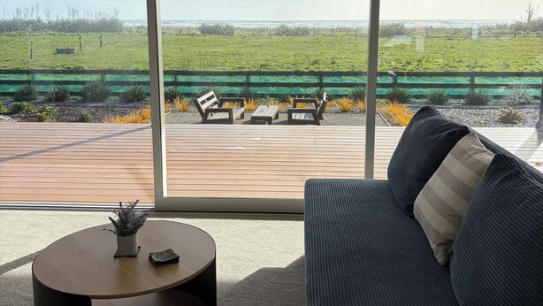 Een moderne woonkamer met een groot raam, een donkere bank, een ronde salontafel en uitzicht op een grasveld en strand.