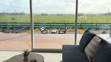 Un salon moderne doté d’une grande fenêtre, d’un canapé foncé, d’une table basse ronde et offrant une vue sur un champ verdoyant et une plage.