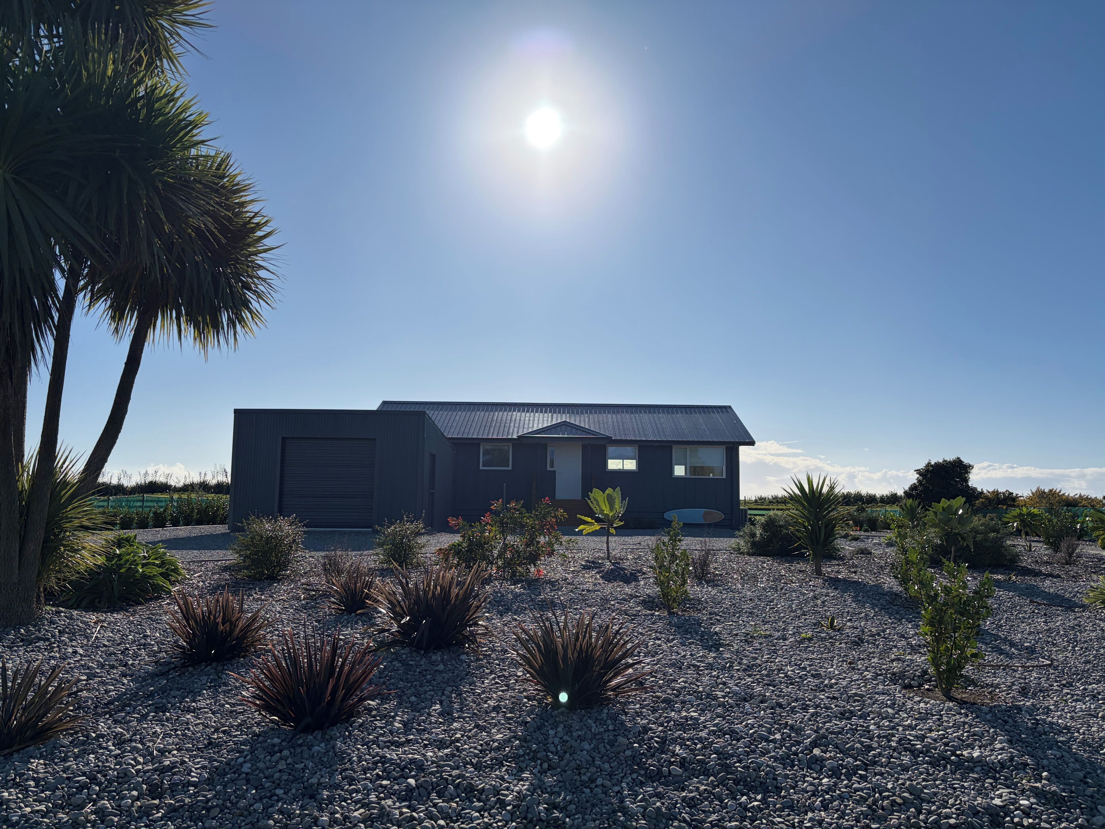 Un bâtiment moderne à deux étages, doté d’un grand portail de garage, entouré d’une allée en gravier et de différentes plantes désertiques.