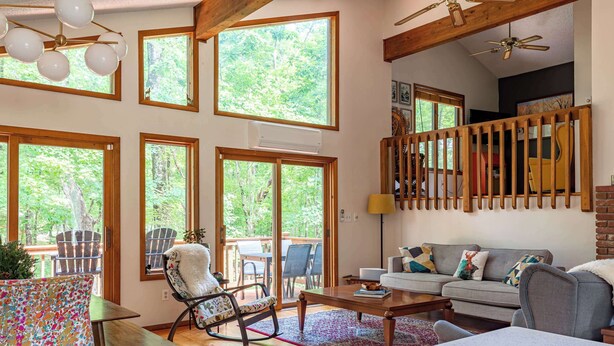 A spacious living room with large windows, a wooden ceiling, a sofa, a rocking chair, and a dining area.