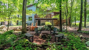 A cabin with a deck, surrounded by trees and a stone fire pit area.
