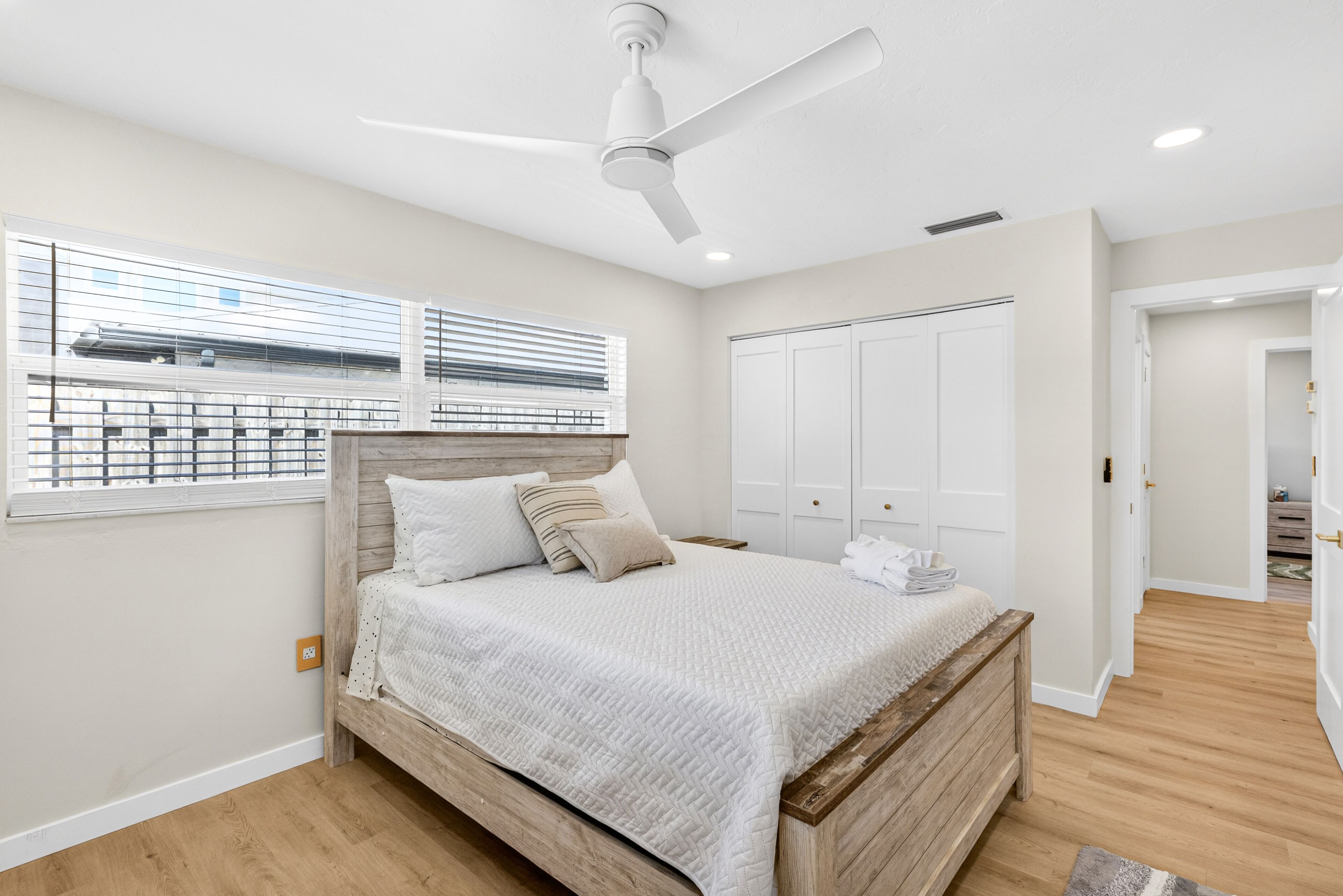 Un dormitorio con cama, ventilador de techo, ventana con persianas y puerta que da a otra habitación.