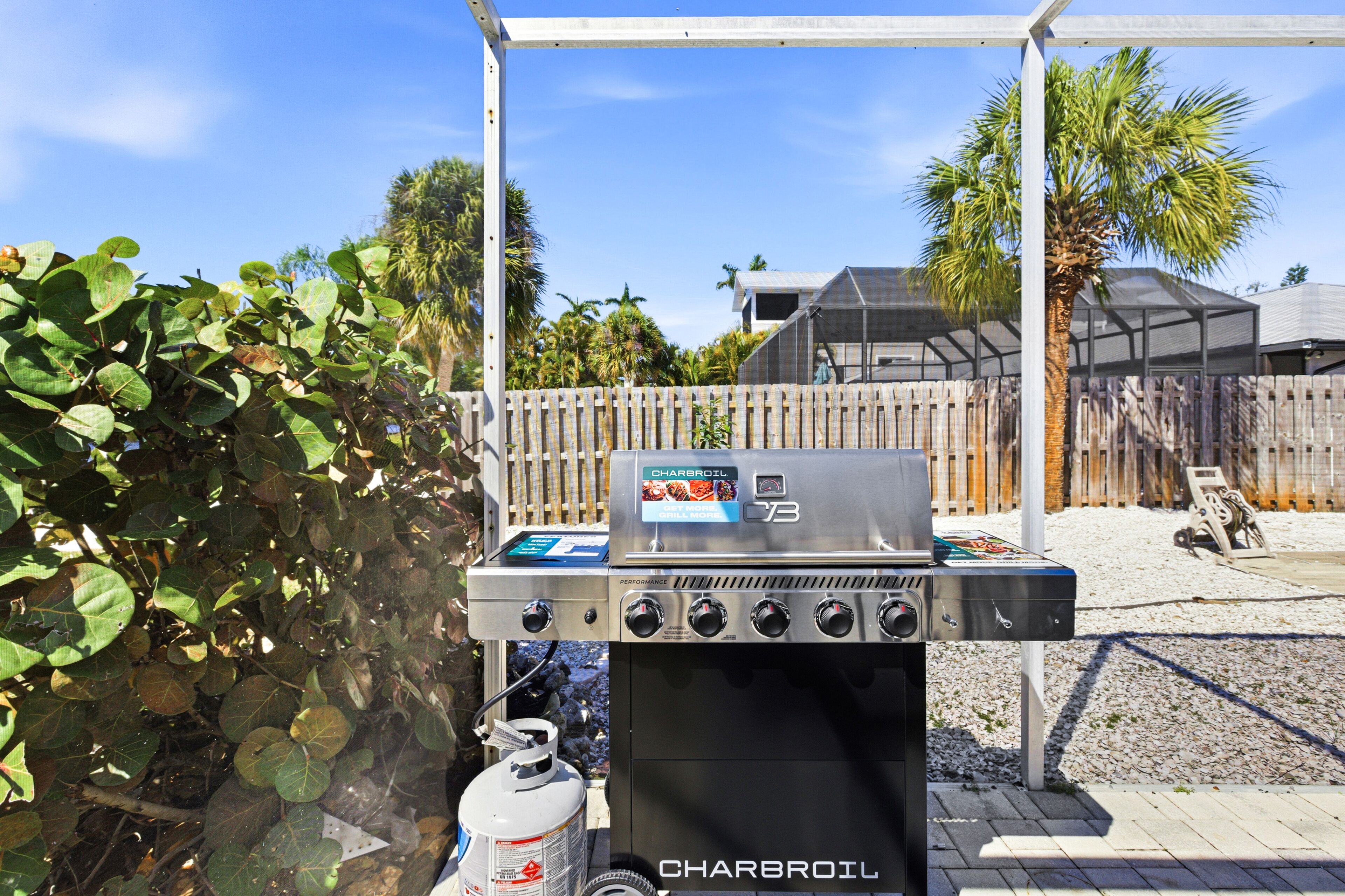A Charbroil outdoor grill with a propane tank, set against a backdrop of a wooden fence, palm trees, and a clear sky.