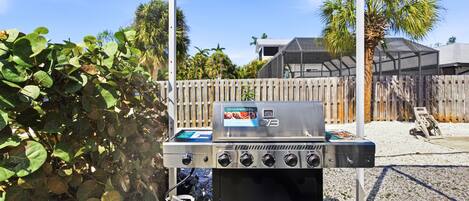 A Charbroil outdoor grill with a propane tank, set against a backdrop of a wooden fence, palm trees, and a clear sky.