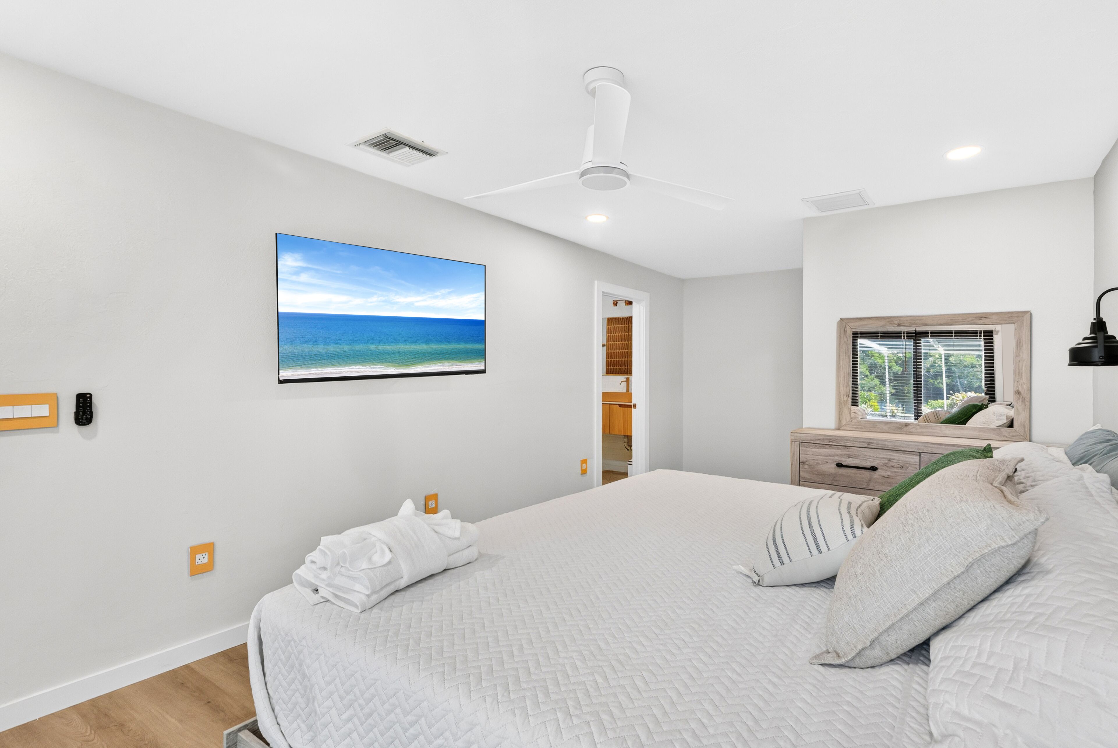 A bedroom with a bed, a ceiling fan, a window with blinds, and a wall-mounted TV displaying a beach scene.