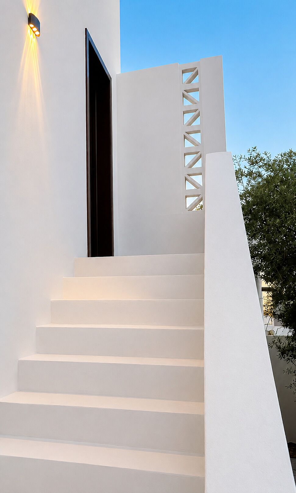 Un escalier blanc orné d’un motif géométrique découpé dans le mur, menant à une porte sombre.