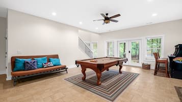 Un vaste espace de vie comprenant une table de billard, un canapé agrémenté de coussins colorés, un ventilateur de plafond et une vue dégagée grâce à de grandes portes-fenêtres coulissantes.