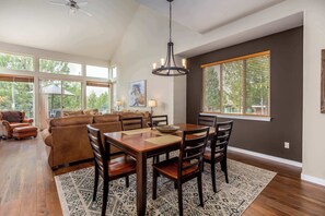 A dining area with a wooden table, chairs, and a chandelier. There is a large window with blinds, a ceiling fan, and a painting on the wall. - Spacious Eagle Crest Condo with Resort Perks (Redmond)