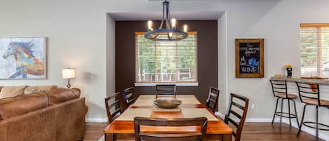 A dining area with a wooden table, chairs, a chandelier, a window with blinds, a couch, and a bar area.