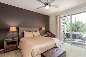 Une chambre avec un lit, deux tables de chevet avec des lampes, un ventilateur de plafond, une grande fenêtre donnant sur les arbres et une porte-fenêtre coulissante qui mène à un patio.