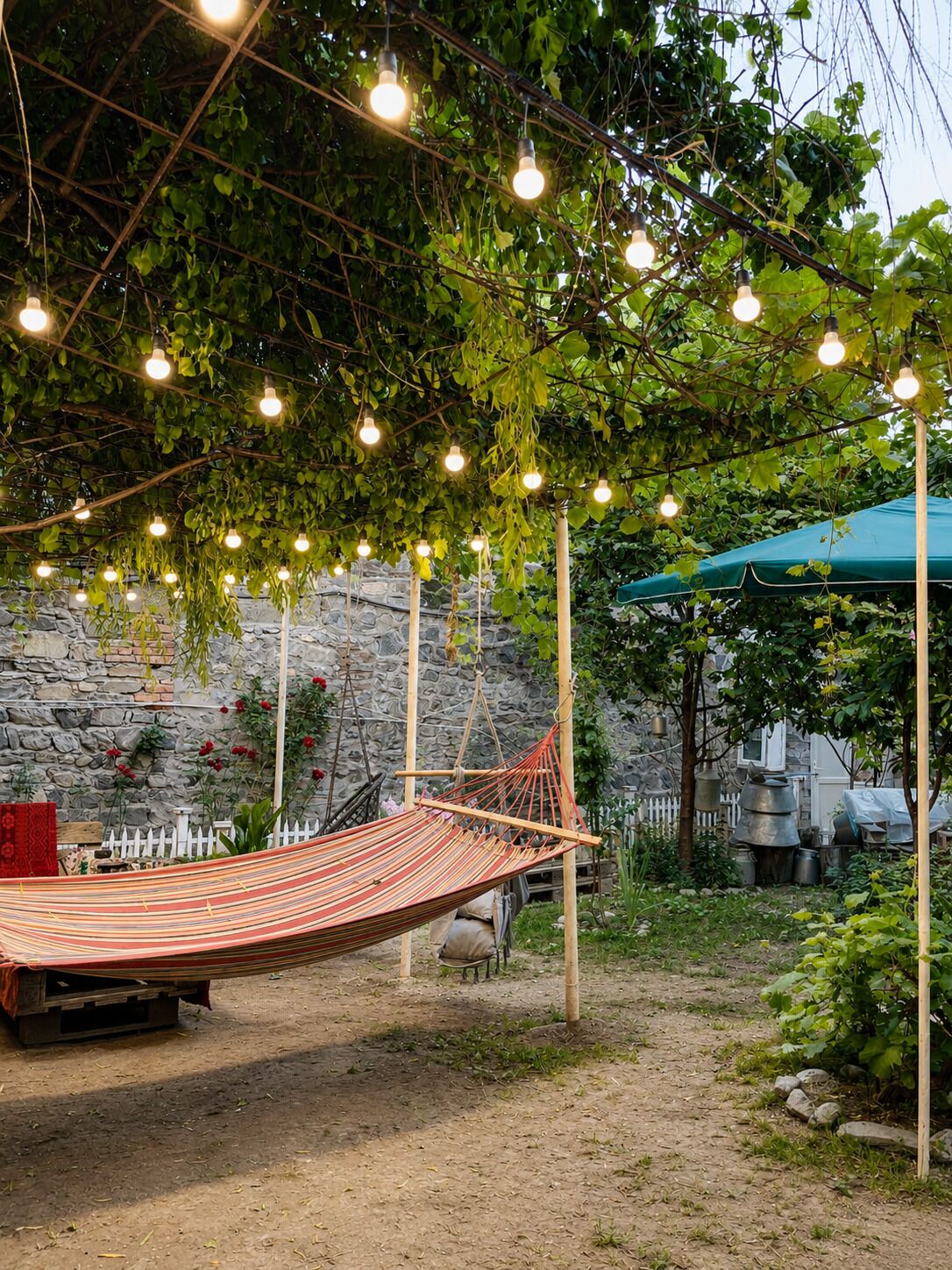 A hammock under string lights, a stone wall, and a green umbrella in a garden setting.