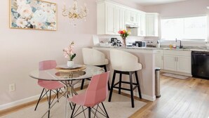 Een keuken met witte kastjes, een eethoek met een glazen tafel en roze stoelen, en een kroonluchter.