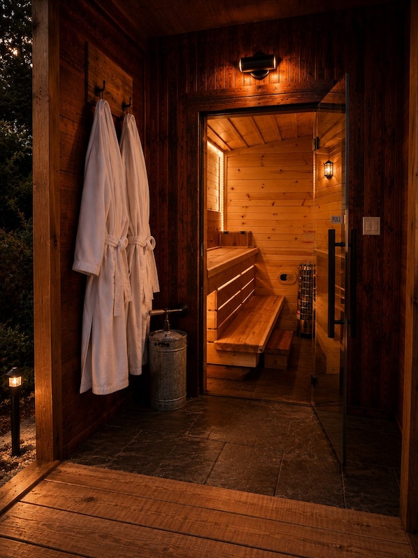 A sauna with wooden walls, benches, and a bench heater. There are towels hanging on the wall and a metal container on the floor.