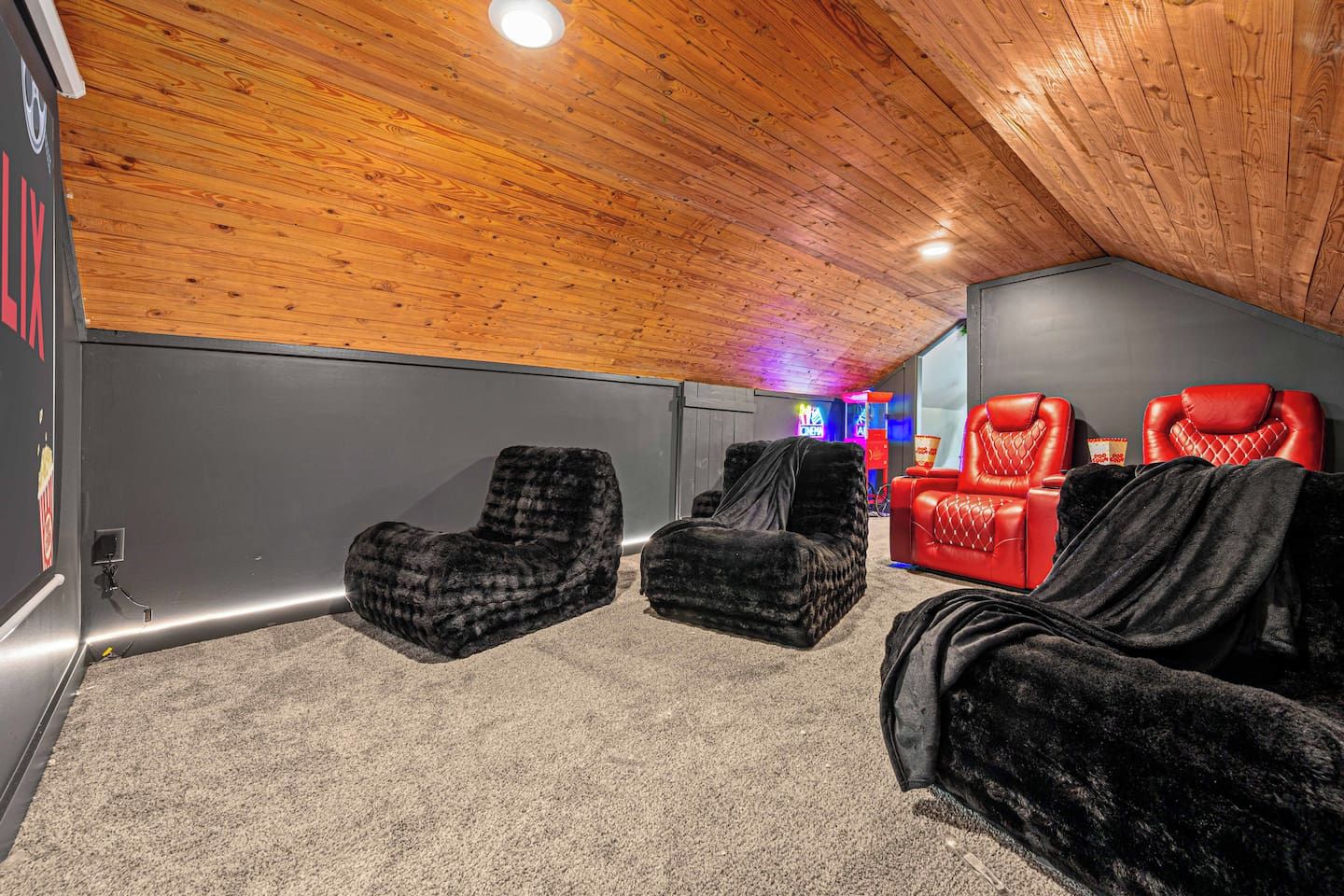 A room with a sloped ceiling, two black cushioned chairs, a red leather chair, and a small table with a neon sign in the background.