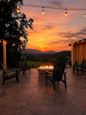 An outdoor seating area with chairs and a fire pit, under string lights, with a sunset view.