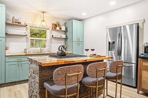 A modern kitchen with a central island, bar stools, a stainless steel refrigerator, and a window with a view of greenery.