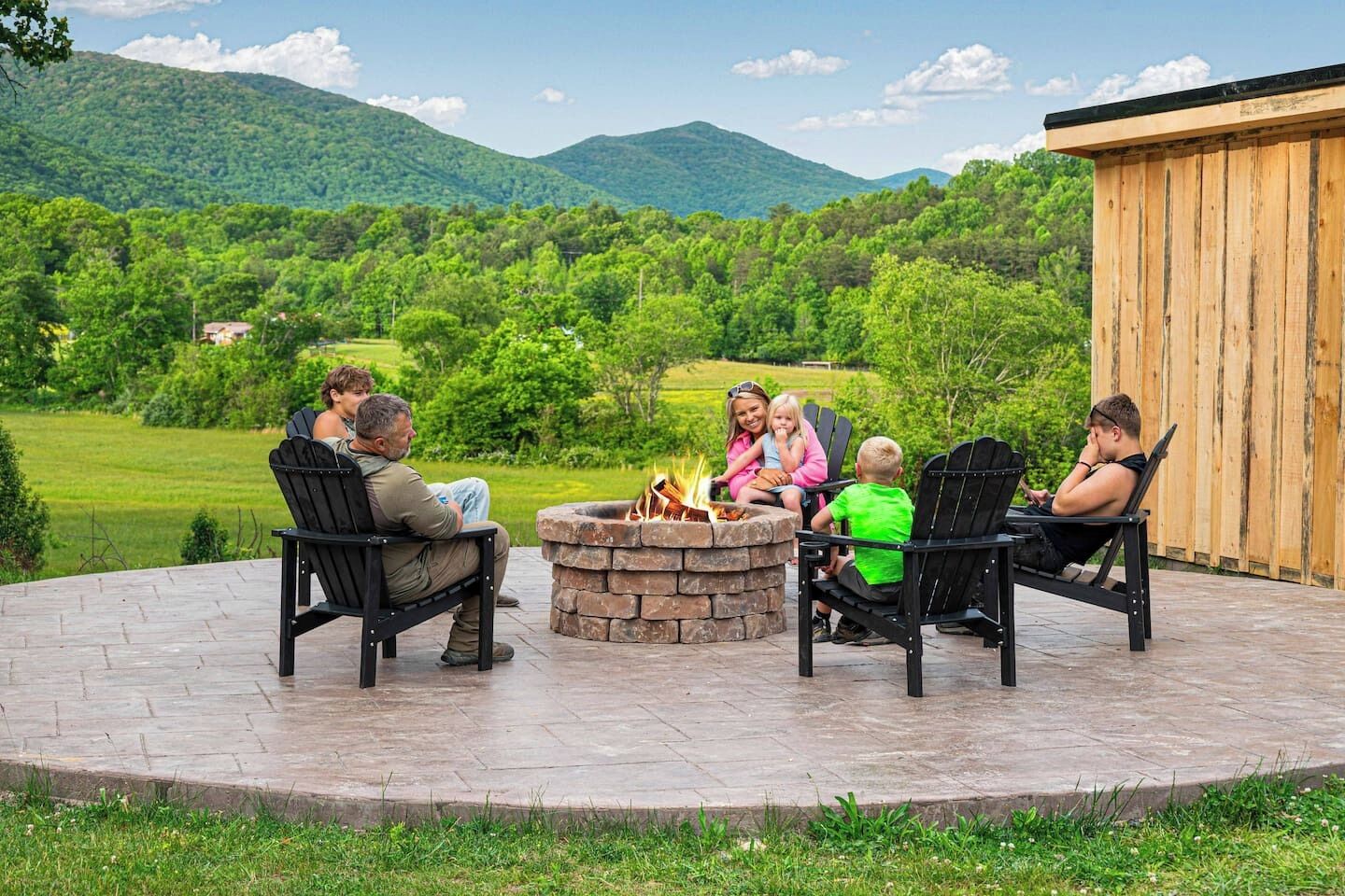 Un grupo de personas sentadas alrededor de un hogar de piedra en un patio con vistas a un paisaje verde y montañas al fondo.