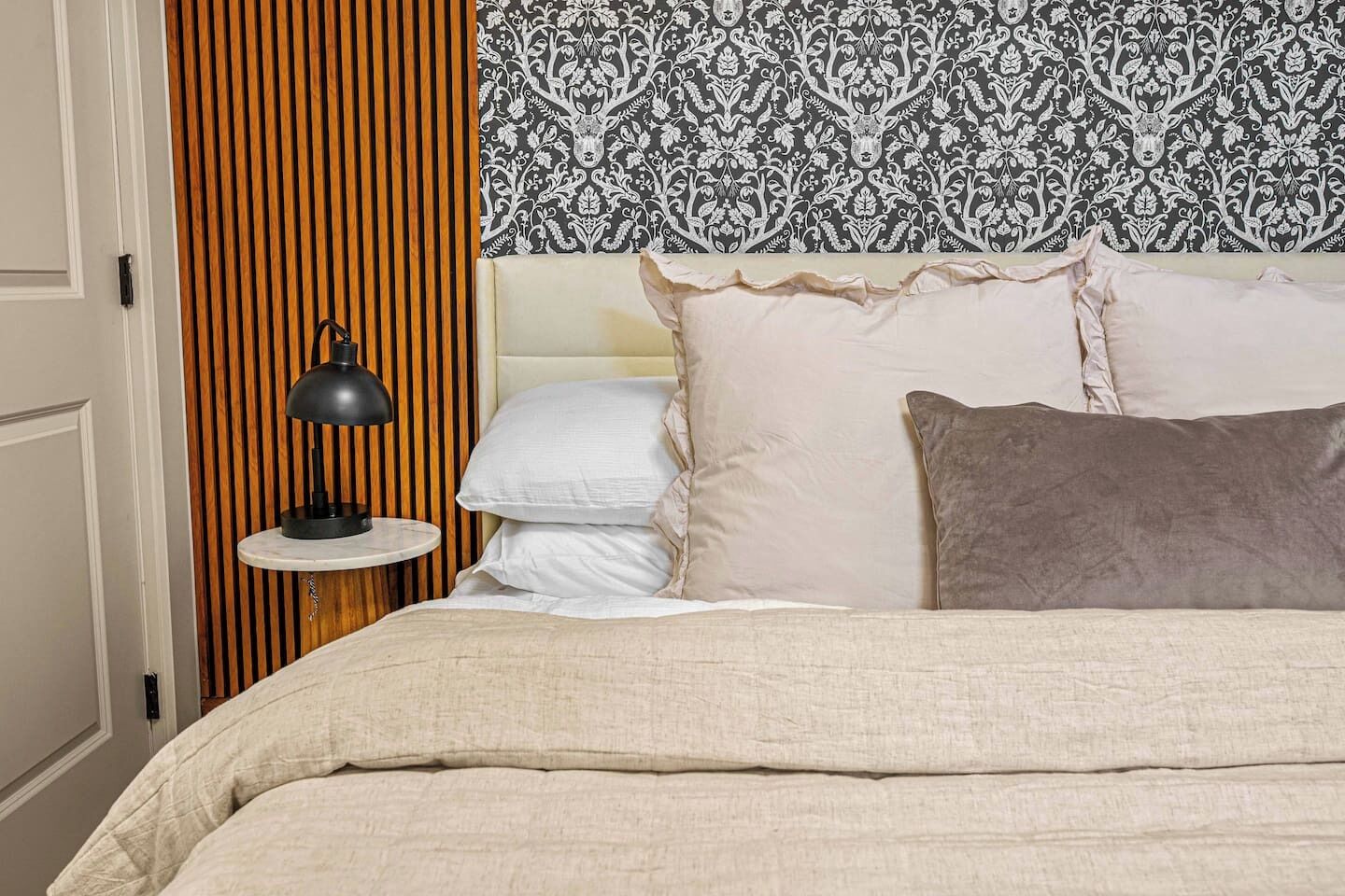 A neatly made bed with a beige comforter and pillows, a bedside lamp on a small table, and a patterned wallpaper on the wall.