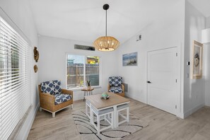 Ein Zimmer mit Esstisch, zwei Korbstühle mit sternförmig gemusterten Kissen, einer Pendelleuchte, einer Tür und einem Fenster mit Jalousien.