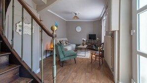 A staircase with a wooden handrail and a brass finial, a green armchair, a white sofa, a small table with a vase, and a television on a stand.