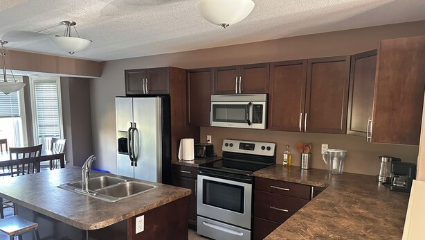 A kitchen with dark wooden cabinets, stainless steel appliances, a granite countertop, and a central island with bar stools. - Modern Duplex with 3 bedroom (Dawson Creek)