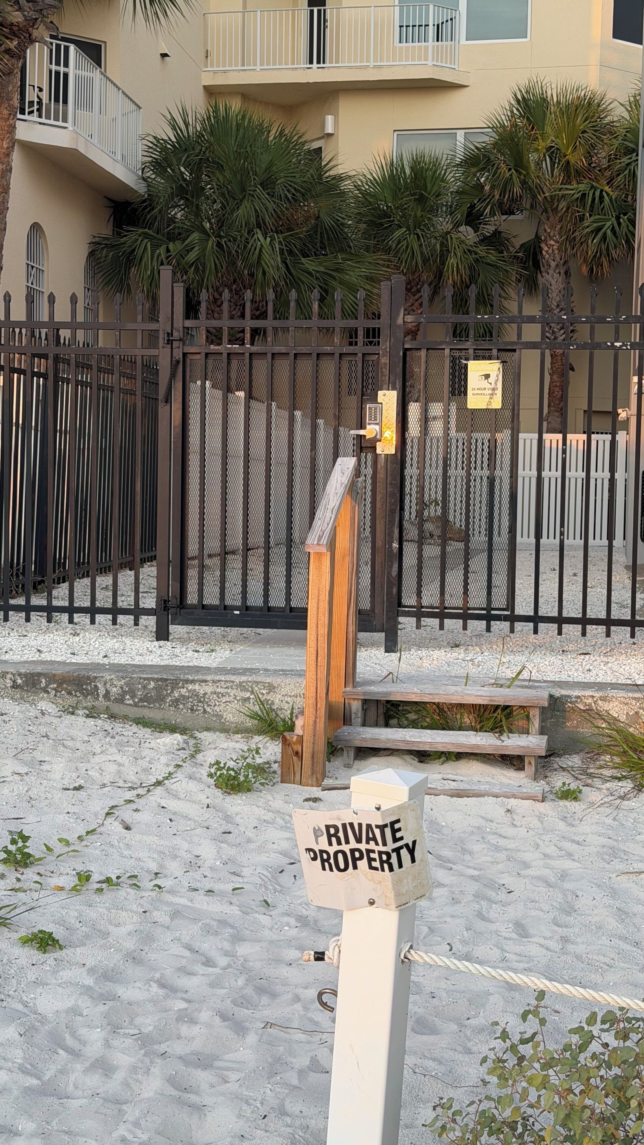 Een toegangspoort met een 'PRIVATE PROPERTY' bord, een metalen hek en een gebouw met balkons en palmbomen op de achtergrond.