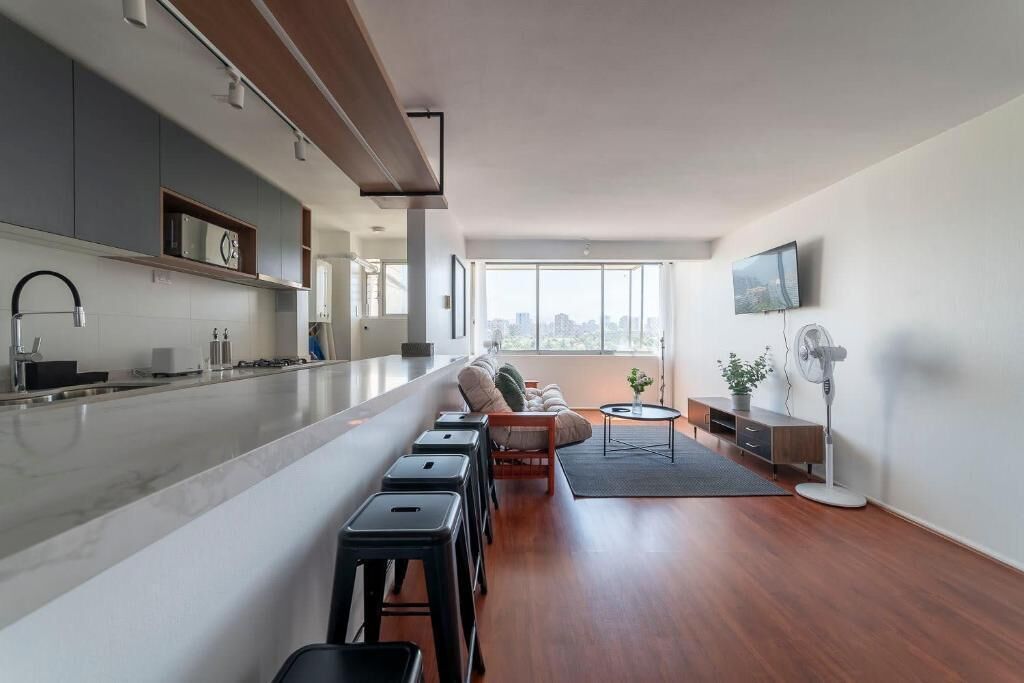 Uma cozinha moderna com uma ilha grande, uma sala de jantar com sofá e banquetas, e uma sala de estar com televisão e ventilador.